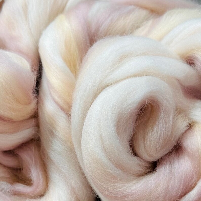 May include: Close-up of a pile of soft, white wool roving with subtle hints of pale pink and yellow. The fibers are tightly packed, creating a textured, cloud-like appearance. Ideal for fiber art projects like spinning and felting.