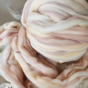 May include: Close-up of a bundle of soft, pastel-colored wool roving. The roving is a mix of white, pale pink, and light yellow, creating a gentle, layered effect. The fibers are loosely spun, suggesting a craft project like spinning or felting.