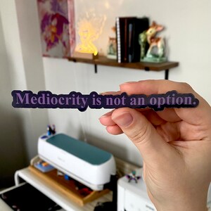Könnte beinhalten: Ein dunkelvioletter Aufkleber mit dem Text "Mediokrität ist keine Option." Der Aufkleber wird von einer Hand gehalten.