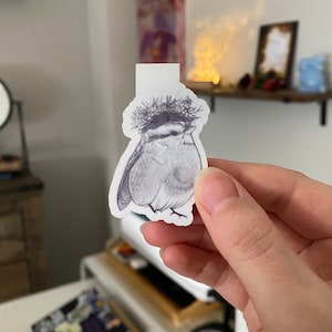 May include: A black and white illustration of a bird wearing a floral crown. The bird is a bookmark with a white tab at the top. The image is held in a person's hand.