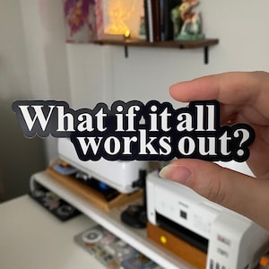 May include: A black and white sticker with the text "What if it all works out?" in a bold, sans-serif font. The sticker is held in a hand, with a blurred background including a printer and shelves.
