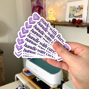 May include: A stack of purple and white stickers with the text "made with flare, handle with care" repeated on each sticker. The stickers are heart-shaped and have a small plus sign in the top corner.