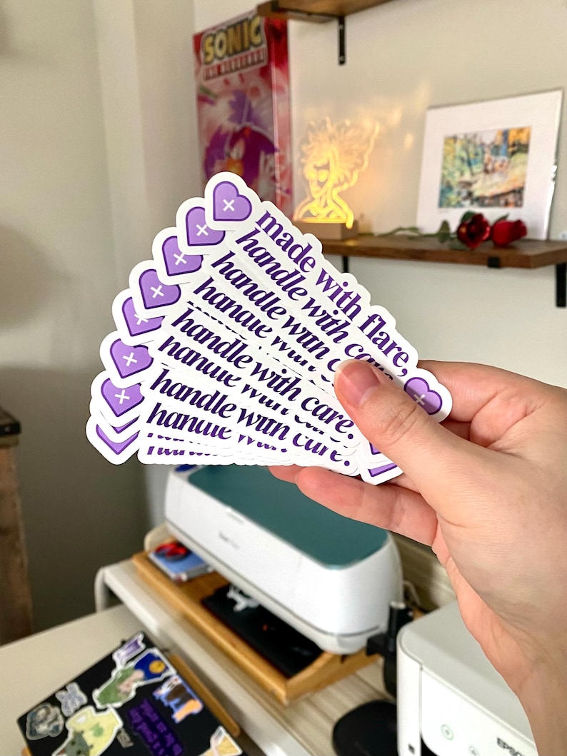 May include: A stack of purple and white stickers with the text "made with flare, handle with care" repeated on each sticker. The stickers are heart-shaped and have a small plus sign in the top corner.