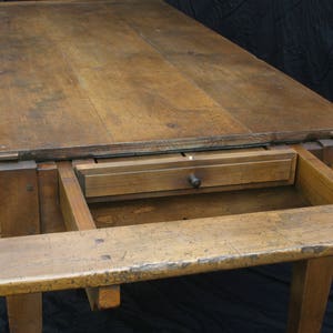 Puede incluir: Una mesa de madera con una extensi&oacute;n de hoja abatible y un caj&oacute;n. La mesa est&aacute; hecha de madera natural con un acabado r&uacute;stico. La mesa est&aacute; en una habitaci&oacute;n oscura con un fondo negro.