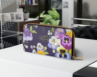 Op de afbeelding: Een donkerpaarse portemonnee met een bloemenpatroon met paarse, gele en witte viooltjes. De portemonnee heeft een gouden rits en is gemaakt van duurzaam materiaal. Een kleurrijk en elegant accessoire.