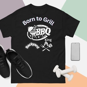 May include: Black t-shirt with a white graphic that reads "Born to Grill BBQ King Backyard." The design includes a grill, stars, and crossed grilling tools. A pair of black sneakers and white dumbbells are visible.