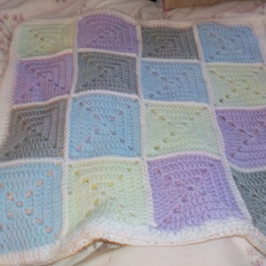 Puede incluir: Una manta de bebé de ganchillo con un diseño de patchwork en tonos de azul, verde y morado. La manta está hecha de hilo suave y tiene un borde blanco.