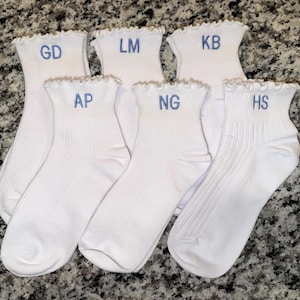 Könnte beinhalten: Eine Auswahl weißer Knöchelsocken mit Rüschen. Jede Socke ist mit blauen Initialen bestickt: GD, LM, KB, AP, NG und HS. Die Socken sind auf einer gesprenkelten Oberfläche angeordnet.