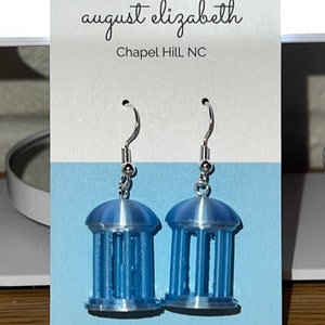 May include: A pair of light blue earrings shaped like miniature columns. The earrings have silver-colored hooks and are displayed on a light blue background. The card behind the earrings reads "august elizabeth Chapel Hill, NC."