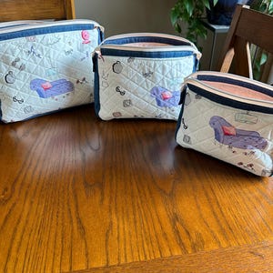 May include: Three quilted cosmetic bags with a white background and a repeating pattern of small drawings and a purple couch. The bags have a denim blue trim and a pink interior. The bags are on a wooden table.
