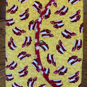 May include: A yellow fabric journal cover with a red border and a repeating pattern of ruby slippers. The cover has a flap that closes with a button.
