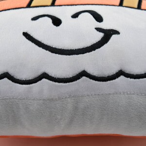 May include: A plush pillow shaped like a sushi roll with a smiling face embroidered on it. The pillow is light gray with black embroidery and has a light orange bottom.