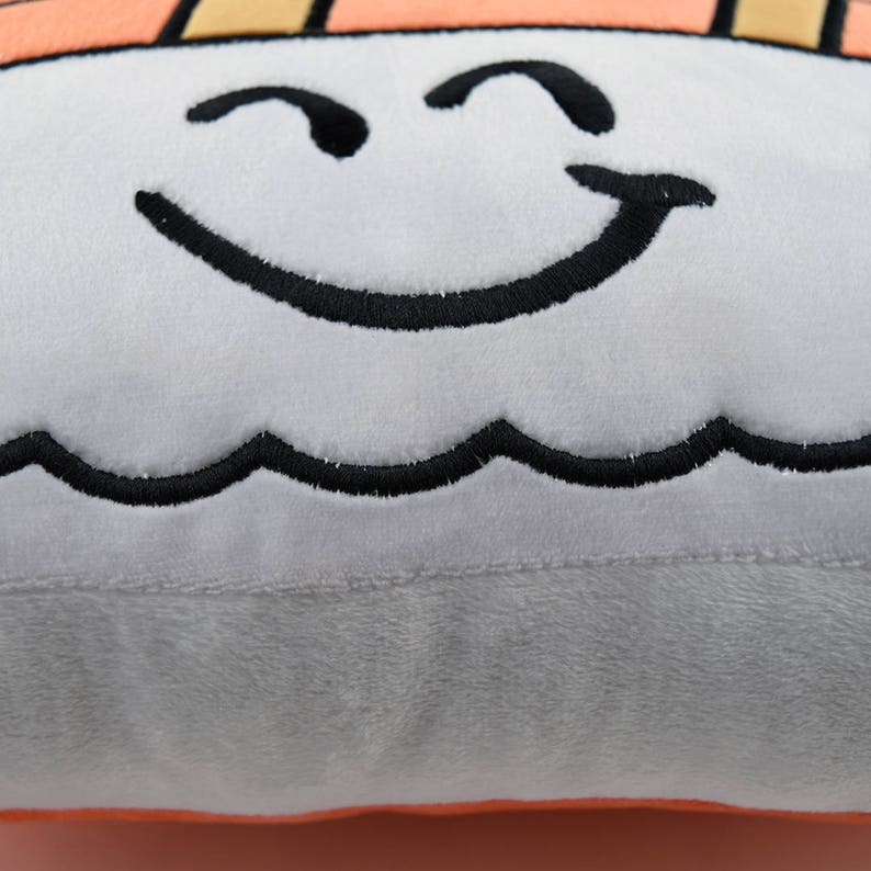 May include: A plush pillow shaped like a sushi roll with a smiling face embroidered on it. The pillow is light gray with black embroidery and has a light orange bottom.