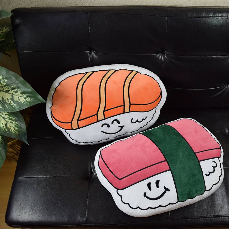 May include: Two plush pillows shaped like sushi. One is a salmon nigiri with a smiling face and the other is a pink tuna maki with a green seaweed wrap and a smiling face.