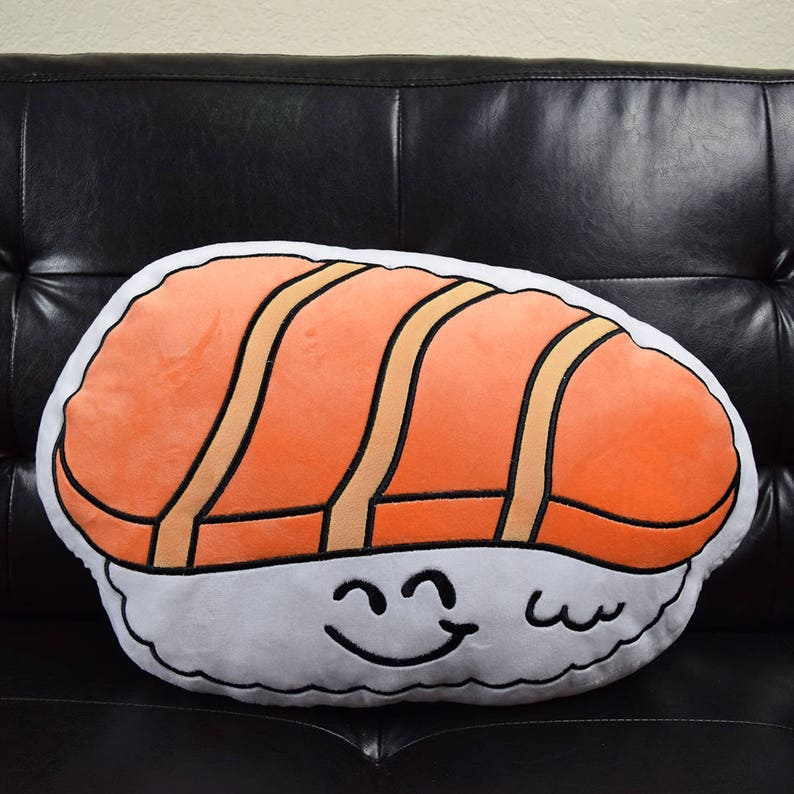 May include: A plush salmon sushi pillow with a smiling face. The pillow is orange and white with black outlines.