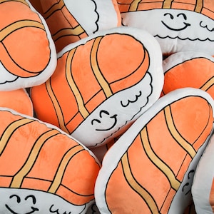 May include: A group of plush pillows shaped like sushi rolls with salmon filling. The pillows are white with orange salmon and black lines. Each pillow has a smiling face with a black outline.