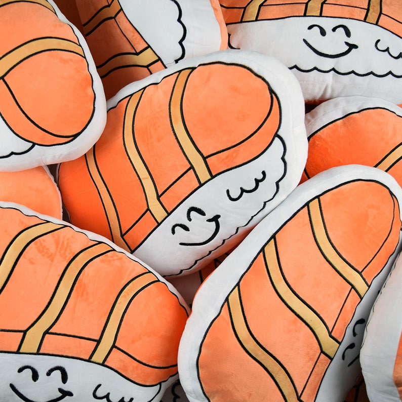 May include: A group of plush pillows shaped like sushi rolls with salmon filling. The pillows are white with orange salmon and black lines. Each pillow has a smiling face with a black outline.