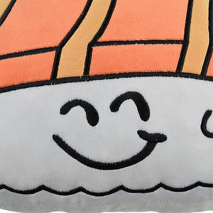 May include: A gray plush pillow shaped like a sushi roll with a smiling face. The sushi roll is orange and yellow with black outlines.