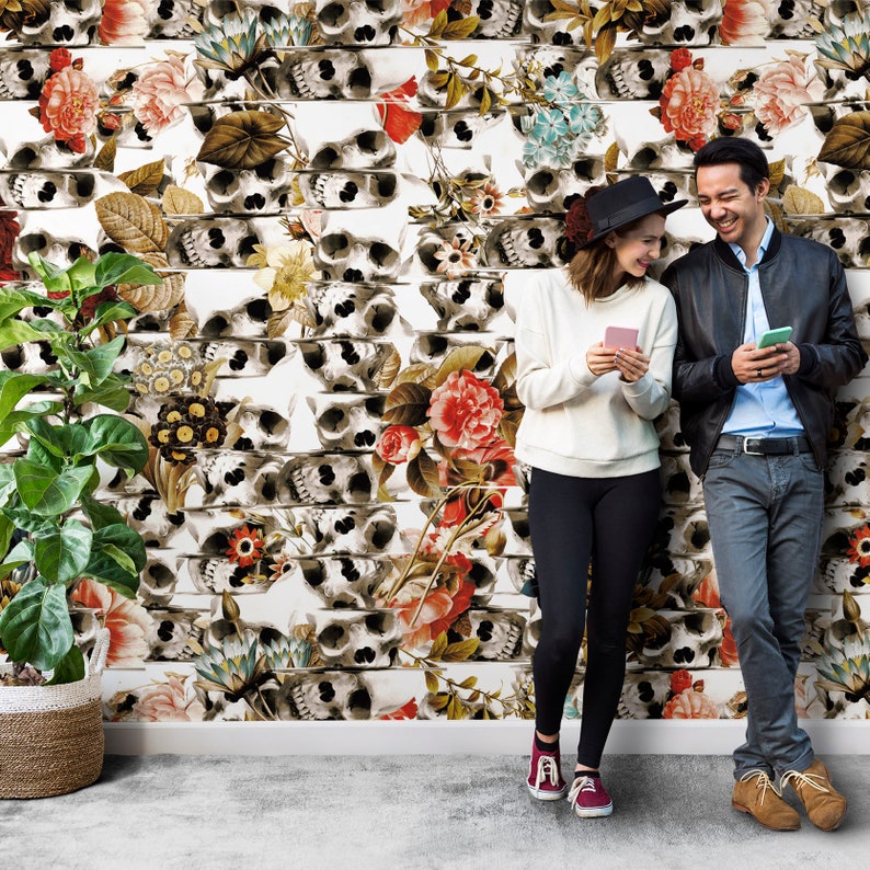 K&ouml;nnte beinhalten: Eine wei&szlig;e Wand mit einem sich wiederholenden Muster aus Totenk&ouml;pfen und Blumen in Rot-, Rosa- und Gr&uuml;nt&ouml;nen. Links steht eine Topfpflanze in einem geflochtenen Korb. Ein Paar steht vor der Wand und h&auml;lt jeweils ein Smartphone.