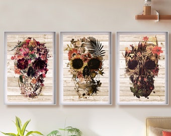 May include: Set of three framed art prints featuring floral skull designs. Each skull is composed of various flowers and foliage in shades of red, purple, and yellow, set against a light wood-grain background. The frames are white.