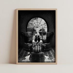 May include: A framed black and white art print depicting two black cats sitting on a small round table covered with a patterned cloth. The table is positioned between two high-backed chairs in front of a large arched window with a view of trees.