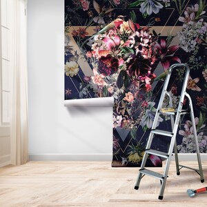 May include: A roll of floral wallpaper with a dark navy background and a geometric design is partially installed on a white wall. The wallpaper features a variety of flowers in shades of pink, purple, and yellow. A silver step ladder is positioned next to the wallpaper.