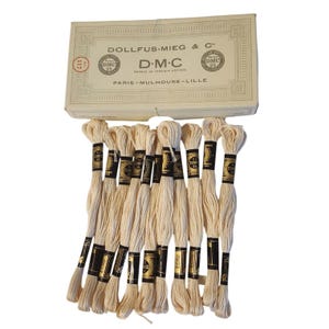 May include: A collection of off-white embroidery floss skeins arranged below a rectangular cardboard box labeled Dollfus-Mieg & Cie D.M.C. The box features black text and circular logos, while each individual skein has a black paper band.