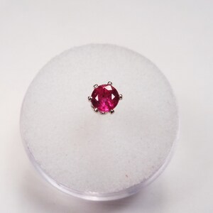 May include: A close-up of a small, round, red gemstone nose ring. The ring is set in a silver setting and is displayed on a white background.