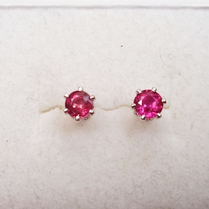 May include: A pair of stud earrings featuring round, vibrant pink gemstones set in silver-colored metal. The earrings are displayed in a white jewelry box, showcasing their bright color and simple design. These earrings are a classic jewelry item.