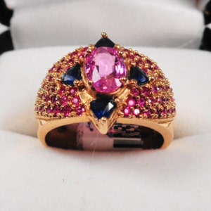 K&ouml;nnte beinhalten: Ein goldener Ring mit einem gro&szlig;en ovalen rosafarbenen Edelstein, umgeben von kleineren rosafarbenen und blauen Edelsteinen.