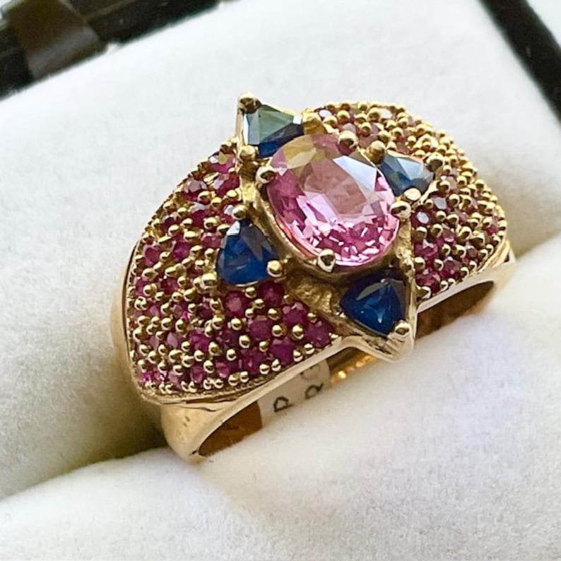 K&ouml;nnte beinhalten: Ein goldener Ring mit einem gro&szlig;en ovalen rosafarbenen Edelstein, umgeben von kleineren rosafarbenen und blauen Edelsteinen. Der Ring ist mit einer Reihe winziger rosafarbener Edelsteine besetzt.