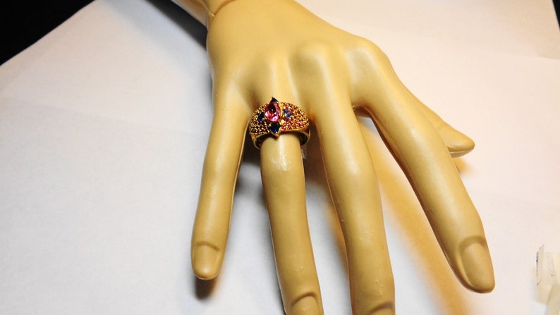 K&ouml;nnte beinhalten: Ein goldener Ring mit einem gro&szlig;en rosafarbenen und blauen Edelstein, umgeben von kleineren blauen Edelsteinen.