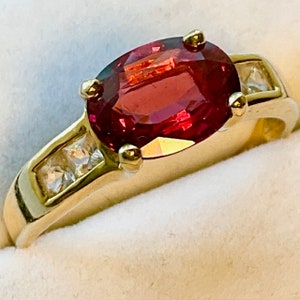 May include: A gold ring with a large oval red gemstone surrounded by smaller clear gemstones.