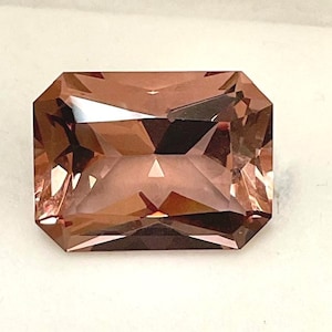 May include: A faceted, rectangular gemstone with a warm, reddish-brown hue. The gem has a step-cut design, with multiple facets creating a play of light and shadow. The stone's edges are precisely cut, giving it a polished appearance.