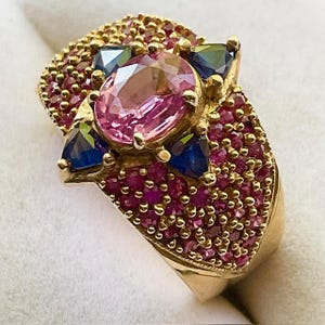 K&ouml;nnte beinhalten: Ein goldener Ring mit einem gro&szlig;en rosafarbenen Edelstein, der von kleineren rosafarbenen und blauen Edelsteinen umgeben ist. Der Ring ist mit einer Reihe winziger rosafarbener Edelsteine besetzt.