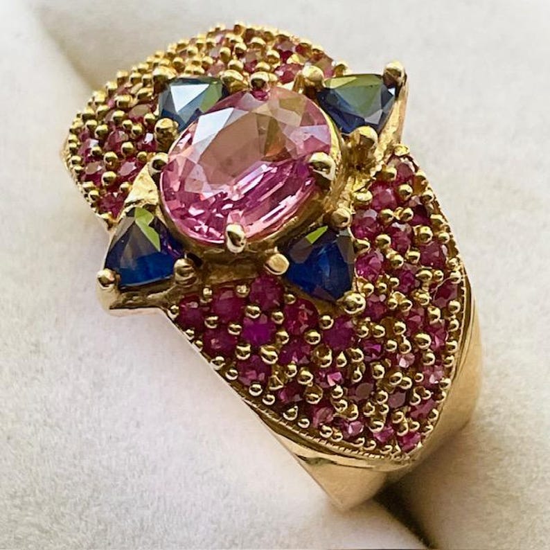 K&ouml;nnte beinhalten: Ein goldener Ring mit einem gro&szlig;en rosafarbenen Edelstein, der von kleineren rosafarbenen und blauen Edelsteinen umgeben ist. Der Ring ist mit einer Reihe winziger rosafarbener Edelsteine besetzt.