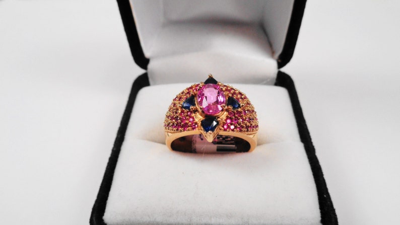 K&ouml;nnte beinhalten: Ein goldener Ring mit einem gro&szlig;en ovalen rosafarbenen Edelstein, umgeben von kleineren rosafarbenen und blauen Edelsteinen.