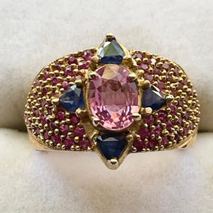 K&ouml;nnte beinhalten: Ein goldener Ring mit einem gro&szlig;en ovalen rosafarbenen Edelstein, umgeben von kleineren blauen Edelsteinen und einer Reihe winziger roter Edelsteine.