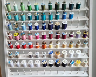 May include: A white wooden thread rack filled with colorful spools of thread. The spools are arranged by color, ranging from greens and blues to pinks, reds, and yellows. The rack has multiple rows of pegs to hold the thread.