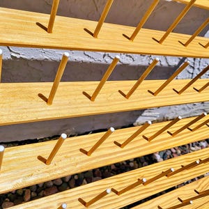 May include: A light brown wooden thread rack featuring multiple horizontal slats with evenly spaced vertical pegs. Each peg is topped with a small white cap, designed for organizing and displaying spools of thread in a structured, orderly arrangement.