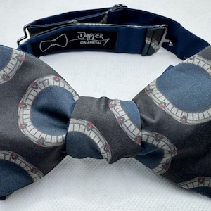 May include: A patterned bow tie with a dark gray and navy blue color scheme. The bow tie features a repeating circular design with red accents. The adjustable neckband is navy blue and has the text "Dapper ON ARRIVAL".