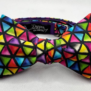 May include: A colorful bow tie with a geometric pattern of triangles in shades of red, orange, yellow, green, blue, purple, and white on a black background. The bow tie is made of fabric and has a purple adjustable strap with the text "Dapper On Arrival".