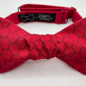 May include: Red bow tie with a dragon scale pattern. The bow tie is made of fabric and has a black adjustable strap with the text "Dapper On Arrival".