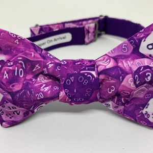 May include: Purple bow tie featuring a pattern of dice. The dice are various shades of purple and white with numbers 1 to 20.