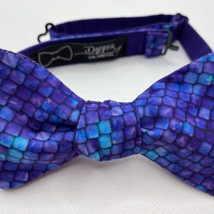 May include: A purple bow tie with a blue and purple scale pattern. The bow tie is made of fabric and has an adjustable strap.