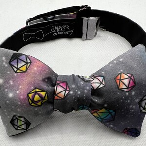 May include: A gray bow tie with a galaxy print and colorful dice. The bow tie has a black adjustable strap with the text "Dapper On Arrival".