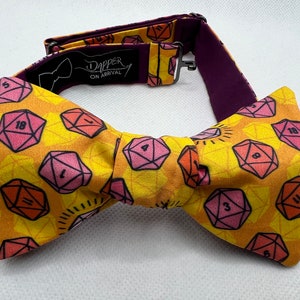 May include: A yellow bow tie featuring a pattern of pink and orange dice with black numbers. The bow tie is tied and has a purple adjustable strap with the text "Dapper On Arrival" in black.