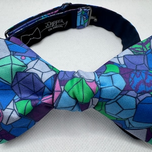 Könnte beinhalten: Eine lebendige Fliege mit einem geometrischen Muster in Blau-, Lila-, Grün- und Rosatönen. Die Fliege hat ein sich wiederholendes Design aus polygonalen Formen. Der Markenname "Dapper On Arrival" ist auf der Innenseite des Nackenbands aufgedruckt.