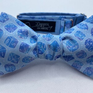 May include: A light blue bow tie with a white background and a pattern of blue dice. The bow tie has an adjustable strap and a black label that reads "Dapper On Arrival".