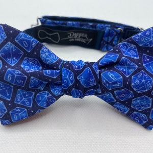 May include: A blue bow tie with a white dice pattern on a navy blue background. The bow tie has a black adjustable strap with the text "Dapper on Arrival" printed on it.